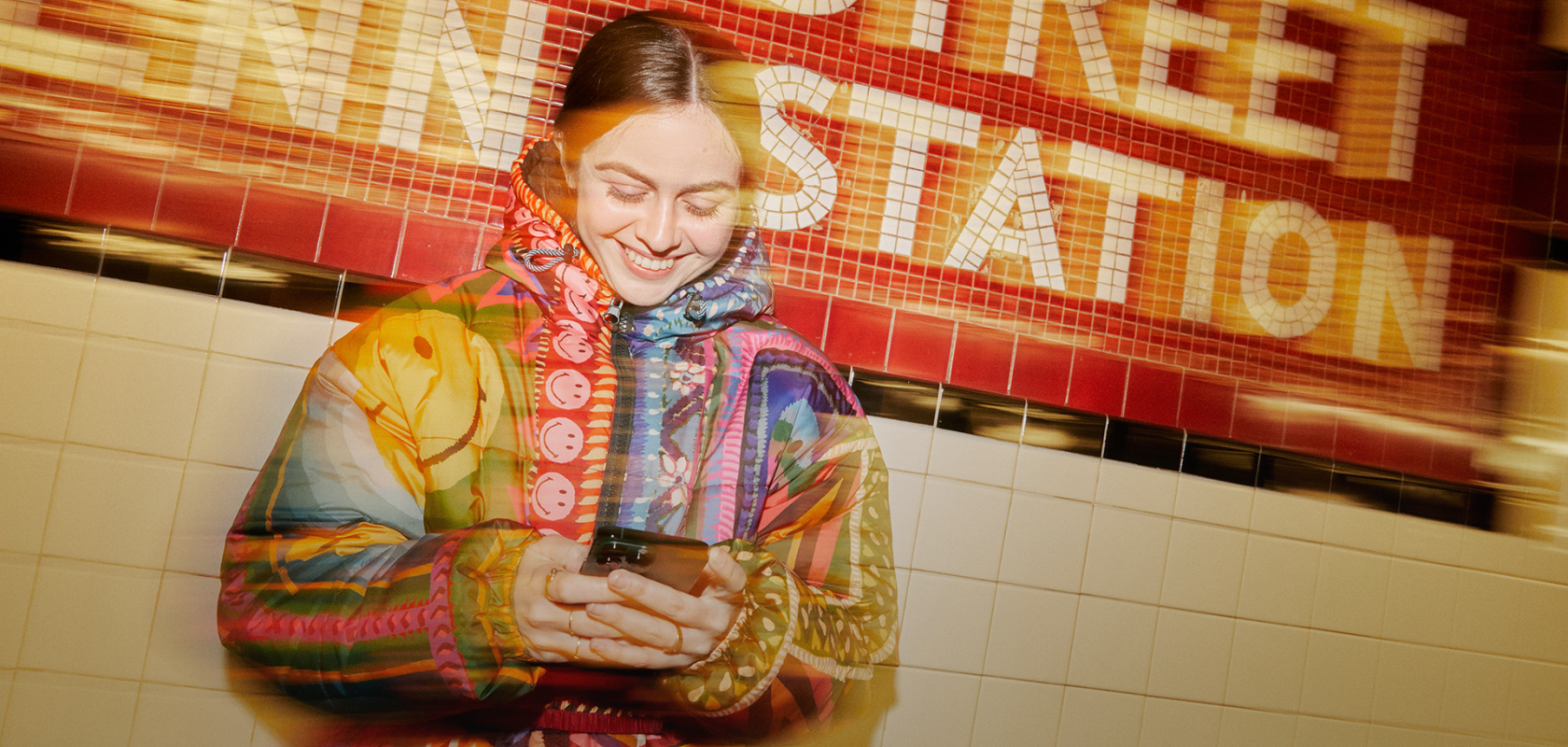 A person smiling while looking at a phone in a subway station, wearing a Smiley x Farm Rio coat.